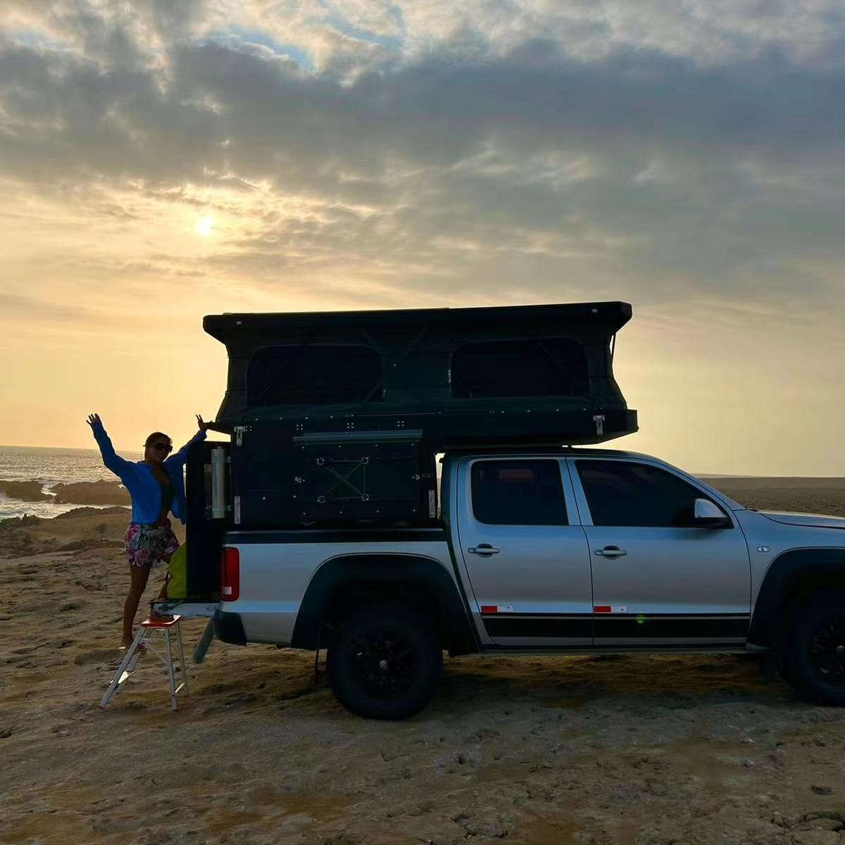 Silver pickup truck fitted with slide in camper shell and rooftop tent at beach sunset, woman with raised arms beside overland camping rig